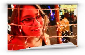 Hispanic young woman with white skin and beautiful smile at a fair of lights at Christmas. Abstract red color on his face. Detail of her hair and big black frame glasses.