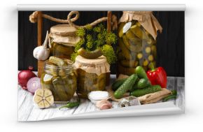 Pickled cucumbers in glass jars and spices and vegetables for preparation of pickles on wooden background.