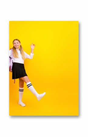 Happy student girl celebrating school success on bright yellow background with backpack and uniform skirt, joyful mood