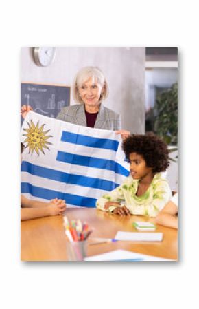 Mature female teacher, conducting a lesson in the classroom, tells the pupils the history of Uruguay and holds the national .flag of the country in her hands