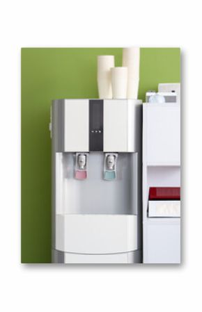 Modern water cooler with paper cups and workplace near green wall