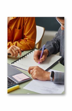 Young adult Caucasian woman and young adult South Asian man studying together at table, writing in notebook, using laptop and reviewing documents in language school classroom setting