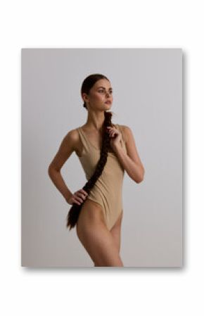 Woman with long braid wearing beige leotard posing on neutral background, showcasing graceful posture and minimalistic style in natural light, studio portrait.