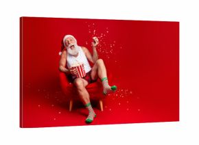 Santa enjoys popcorn on Christmas Eve relaxing in a red chair wearing hat striped socks and smiling