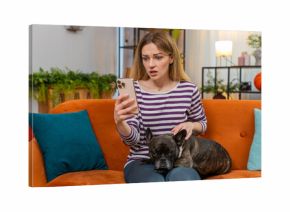 Middle-aged woman with French bulldog at home reads crisis news on smartphone face shows worry sigh and pause. Girl dog on sofa sits near gives comfort warm cuddle loyal friend calm touch caring bond