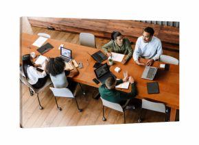 A group of three workers are talking while two female workers are looking at a laptop while sitting at a desk next to them