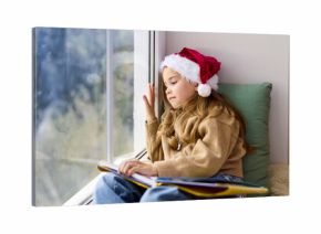 Christmas child in Santa hat reading by window. On a cozy windowsill the child pauses with an open book, touching the window as a Christmas tree glows softly inside. Soft holiday calm at home.