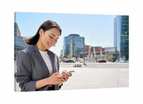 Young happy Asian business woman wearing suit holding mobile phone standing in big city on busy downtown street, smiling lady using smartphone for texting, using apps outside