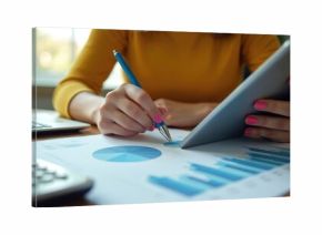 Woman reviews financial charts on tablet and paper. She uses pen to point at data, planning budget with laptop computer nearby. Person analyzes stats for better investment results.