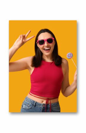 Beautiful young woman in sunglasses with sweet lollipop showing victory gesture on yellow background