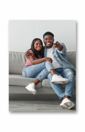 Happy laughing relaxed millennial African American woman and man watching tv in living room interior, holding remote control and hugging. Smiling couple enjoying film and movie evening together