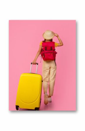 Back view of unrecognizable woman backpacker walking with yellow suitcase on pink studio background, touching her summer hat, full length photo, copy space. Female tourist with backpack and luggage