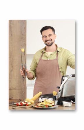 Young happy man cooking fresh corn cobs on modern electric grill in kitchen