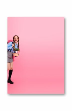 Happy female student holding a plant and boo book s while posing cheerfully against a vibrant pink background