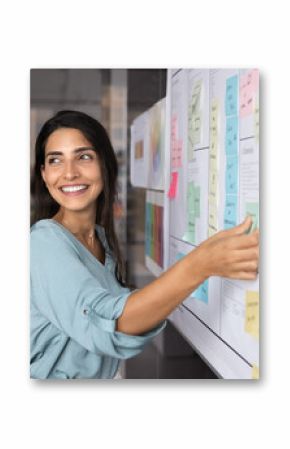Smiling Latina professional woman placing sticky notes on large planning board, engaged in strategic planning, creative brainstorm session or workflow organization. Project management in modern office