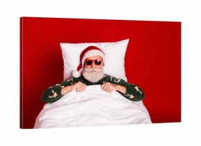 Santa relaxes in a cozy red bedroom wearing a festive pajama sweater and lounging in bed for a playful Christmas photo