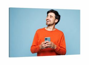 Young fun Caucasian man he wear orange sweatshirt casual clothes hold in hand use mobile cell phone look aside isolated on plain pastel light blue cyan background studio portrait. Lifestyle concept
