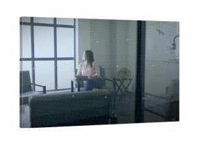 Observing Asian woman sitting thoughtfully in office lounge area, with tablet device and whiteboard