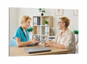 Happy senior patient and nurse on geriatric care appointment in clinic. Young doctor listening to smiling old woman talking about symptoms, general practitioner writing notes on consultation
