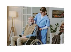 Caucasian senior man with disability sitting in wheelchair being assisted by young adult Hispanic woman caregiver while elderly Caucasian woman reading book in background