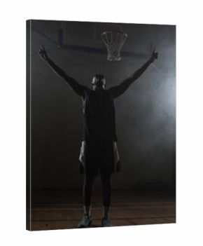 Basketball player standing on wooden court under hoop in dim gym while haze drifts around lights