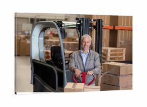 Manual pallet jack carrying stacked cardboard boxes past parked forklift under bright lights