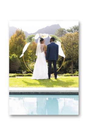 Rear view of african american bride and groom under wedding arch by pool in sunny garden