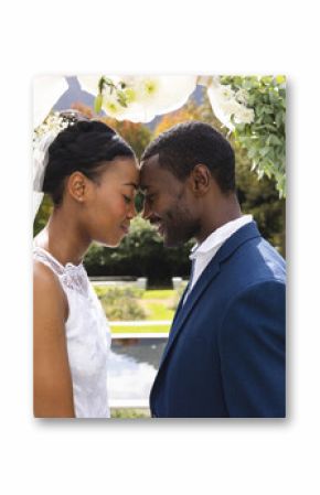 Happy african american bride and groom touching heads under wedding arch at ceremony in sunny garden