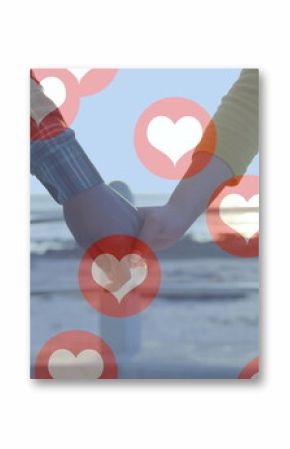 Image of red heart icons floating over mid section of couple in love holding hands by seaside