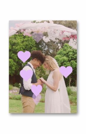 Image of lilac hearts and happy diverse couple holding hands under wedding arch in garden