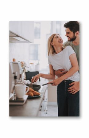 Cooked with love. Cute laughing blonde frying something in skillet. Her bearded husband is hugging her from behind