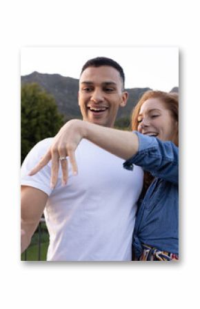 Young couple taking selfie outdoors, showing engagement ring with joyful expressions