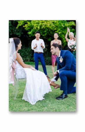 Groom kneeling, removing garter from bride's leg, friends cheering in garden