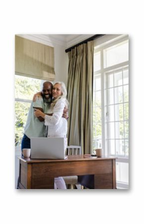Couple embracing joyfully near desk with laptop, sunlight streaming through window