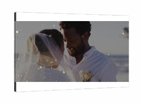 Facing bride and groom standing on sandy beach by ocean, with veil boutonniere and confetti overlay