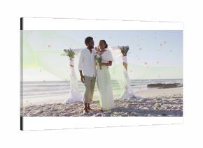 Standing bride and groom wearing wedding attire holding bouquet on sandy beach, with arch backdrop