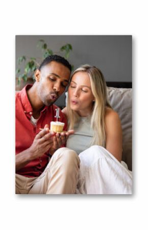 Diverse couple celebrating anniversary blowing candle on cupcake at home, feeling joyful