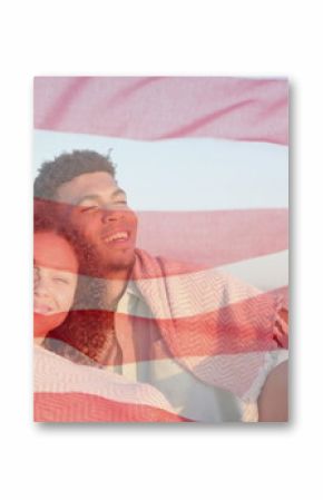 Image of american flag over happy diverse couple sitting on sunny beach embracing