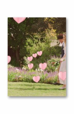 Image of pink hearts over happy diverse couple walking in sunny garden on wedding day