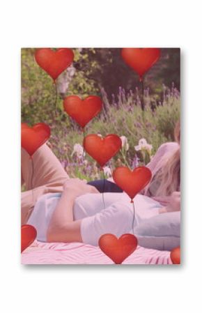 Image of red heart balloons over happy diverse couple relaxing at picnic in sunny garden