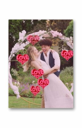 Image of red love you hearts over happy diverse couple embracing in garden on wedding day