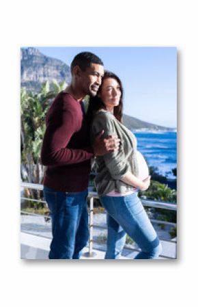 Expecting multiracial couple embracing on balcony with scenic ocean view in background