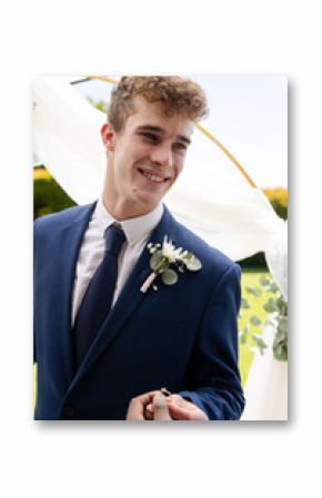 Holding wedding ring, groom smiling during outdoor ceremony with guests watching