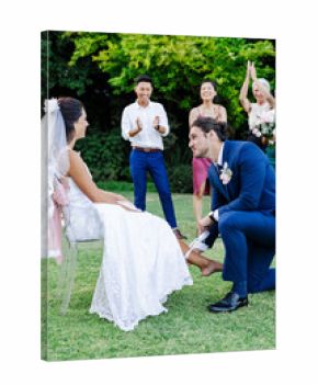 Groom kneeling, removing garter from bride's leg, friends cheering in garden