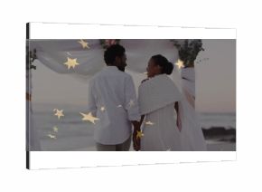 Vowing couple wearing wedding attire on beach at sunset, with draped arch, gold stars, waves