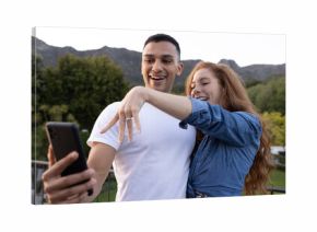 Young couple taking selfie outdoors, showing engagement ring with joyful expressions