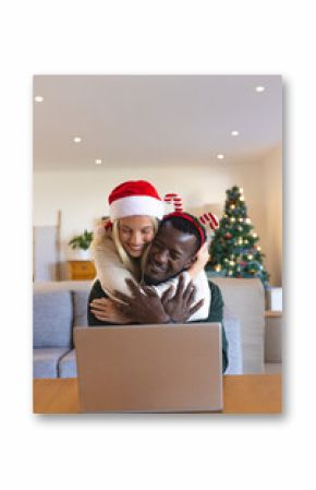 Happy multiracial couple embracing at home with laptop, celebrating Christmas together