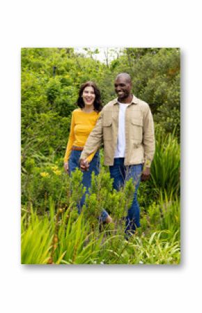 multiracial couple walking hand in hand through lush green garden, smiling happily