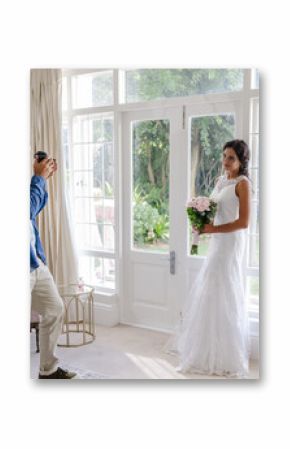 Photographer capturing bride holding bouquet in elegant white dress by window