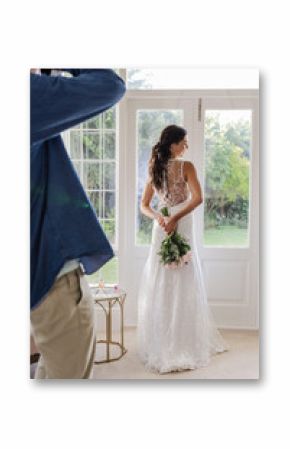 Bride in elegant gown holding bouquet, standing by window, enjoying serene moment
